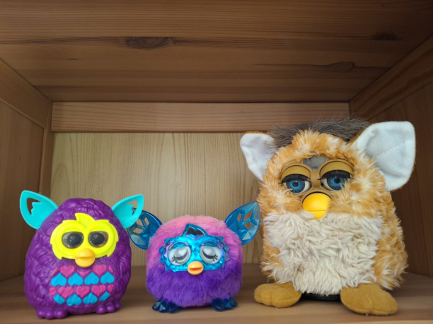 A giraffe coloured original 1998 Furby in reasonable condition with a tiny purple Furbling and similarly-sized purple plastic McDonald's version of a Furby boom!, all sitting on an IKEA shelf.